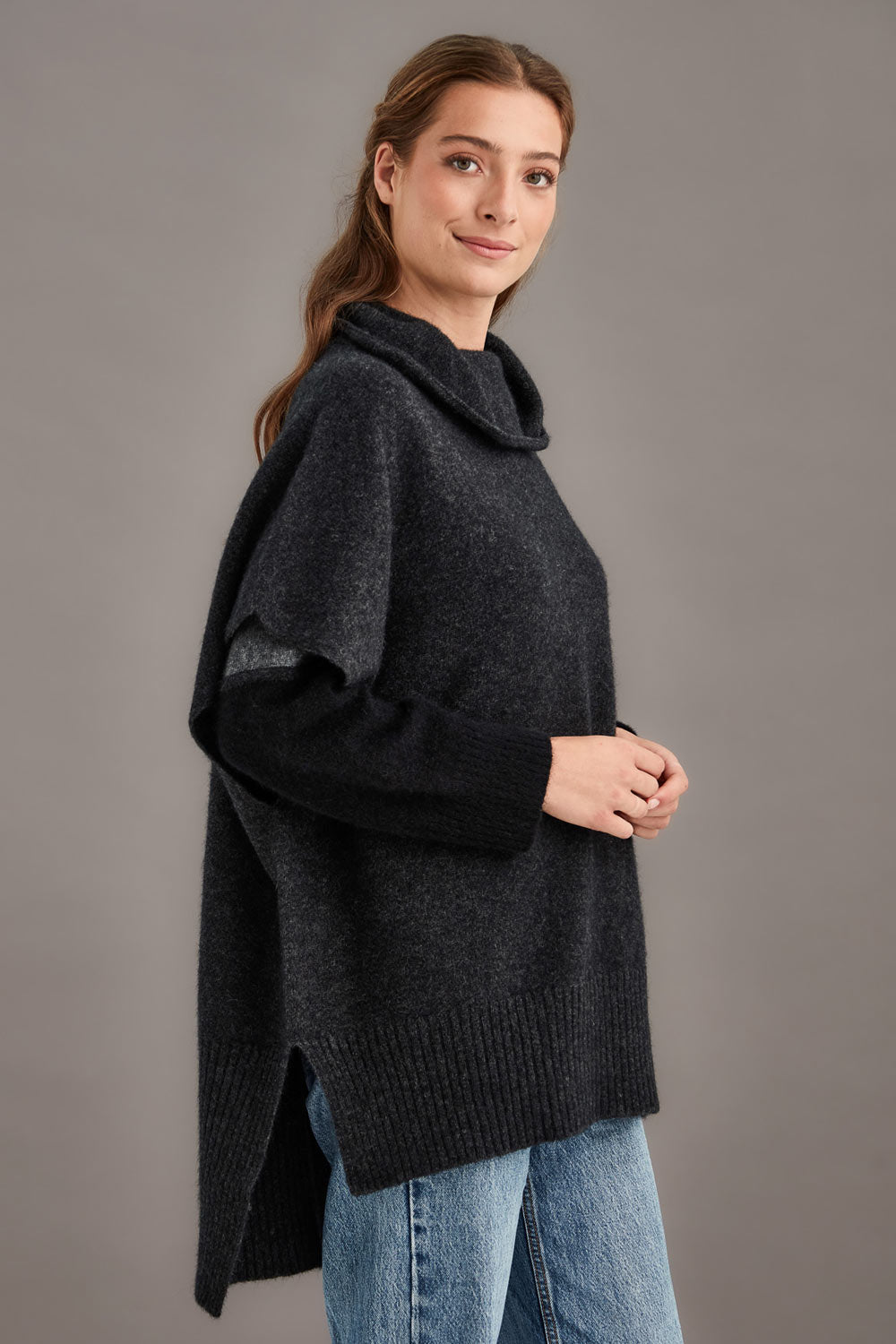 Woman wearing a dark gray poncho over a black top with light blue jeans on a gray background