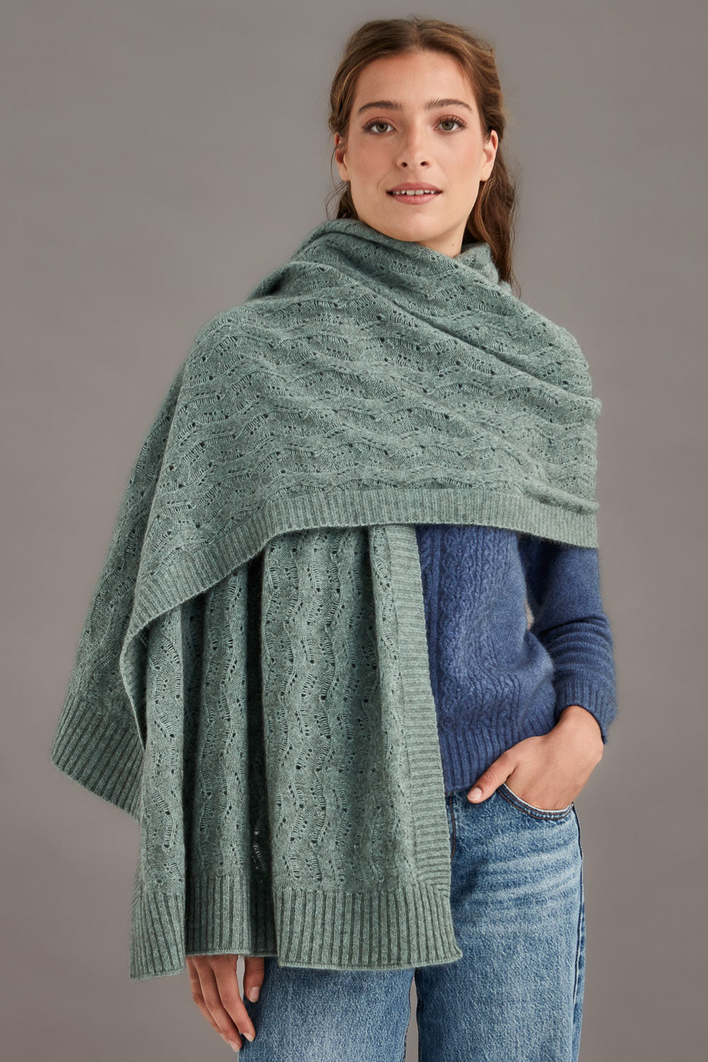 Woman wearing a mint green knitted shawl over a blue sweater against a gray background