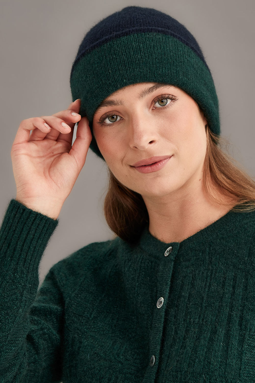 Woman wearing a dark green and navy knitted hat and cardigan against a gray background