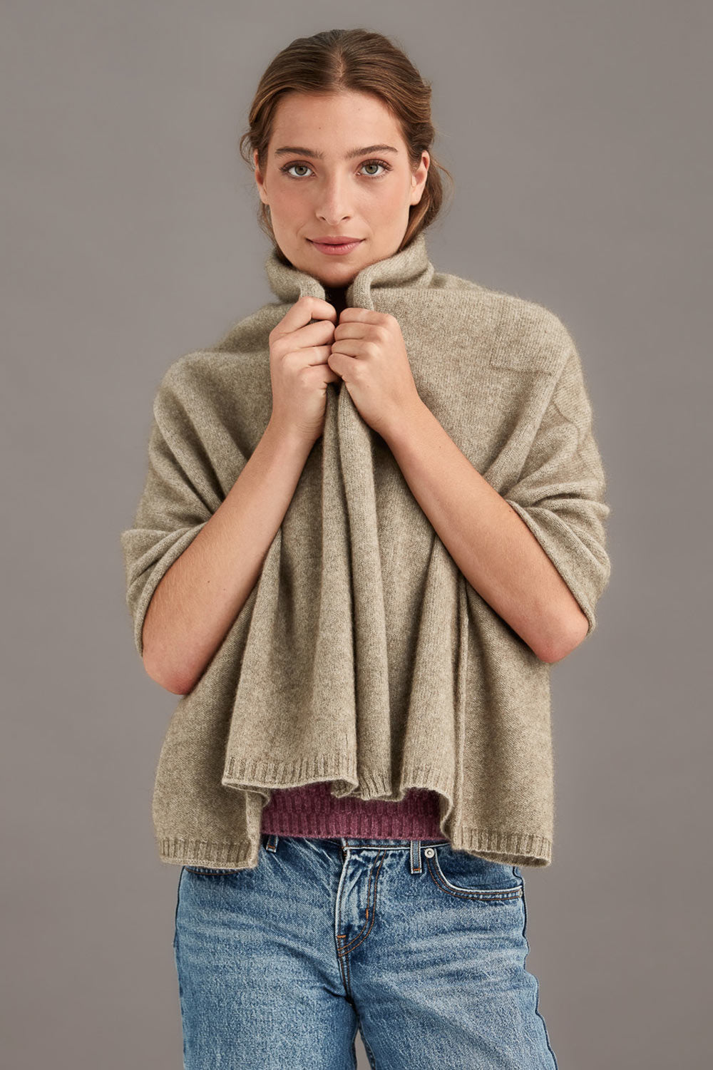 Woman wearing a beige travel throw over a maroon shirt with blue jeans against a gray background